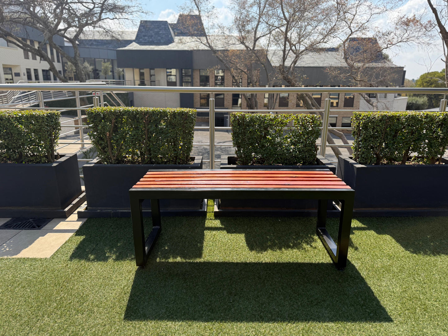 Outdoor setting with a wooden bench on artificial grass, surrounded by green hedges and buildings in the background.