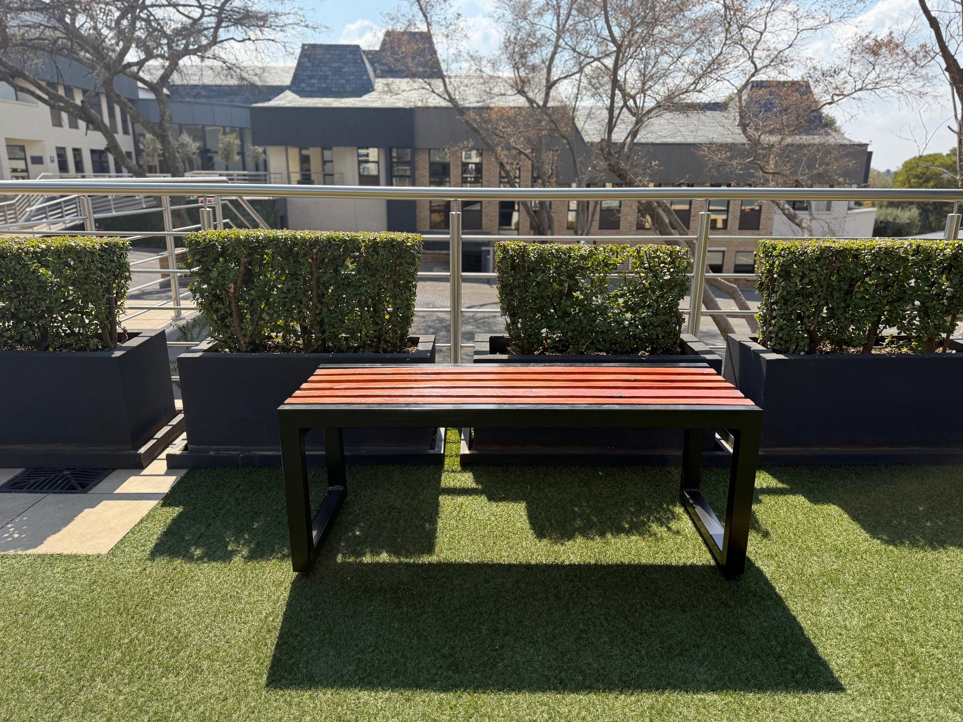 Outdoor setting with a wooden bench on artificial grass, surrounded by green hedges and buildings in the background.